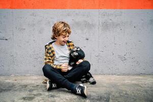 Happy kid sitting near concrete wall and looking at dog photo