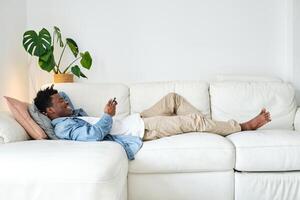 Smiling black man using smartphone and lying on cozy sofa photo