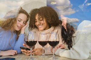 Smiling multiethnic ladies with wine looking at phone screen photo