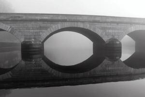 Historic stone bridge shrouded in morning fog reflecting in still water at dawn photo