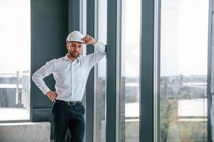 Standing, near panoramic windows. Man in white shirt is working indoors at construction site. Managing the project photo