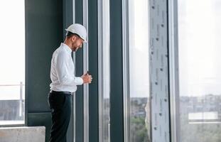 Standing ,near panoramic windows. Man in white shirt is working indoors at construction site. Managing the project photo