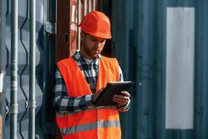 With digital tablet in hands. Male worker is on the location with containers photo