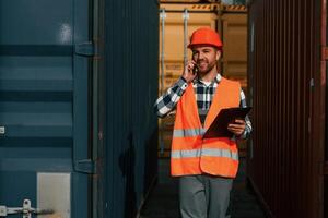 Using phone and ,holding tablet. Male worker is on the location with containers photo