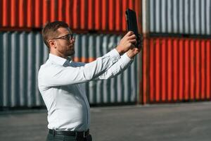 Taking photos by using tablet.. Male worker is on the location with containers