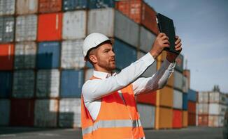 Taking photos, by using tablet. Male worker is on the location with containers photo