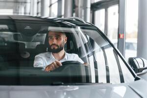 Front view. Sitting ,inside of automobile. Young man in white clothes is in the car dealership photo