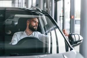 Front, view. Sitting inside of automobile. Young man in white clothes is in the car dealership photo