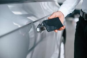 Wit,h smartphone in hand. Young man in white clothes is in the car dealership photo