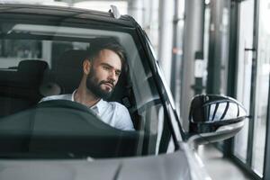 Front view. .Sitting inside of automobile. Young man in white clothes is in the car dealership photo