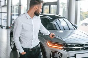 Using the ,keys remote controller. Young man in white clothes is in the car dealership photo