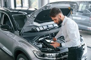 Looking under, the hood and holding tablet. Young man in white clothes is in the car dealership photo