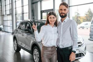 Standing with keys in hands. Happy, owners. Man with woman in white clothes are in the car dealership together photo