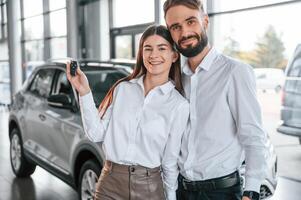 Standing with keys in hands. Happy ,owners.. Man with woman in white clothes are in the car dealership together photo