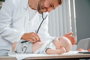 Close ,up view. Using stethoscope. Doctor in white coat is taking care of little toddler indoors photo