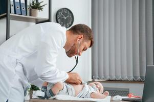 Using ,stethoscope. Doctor in white coat is taking care of little toddler indoors photo