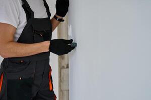 Construction worker in gloves uses laser distance measurer to inspect wall. Builder using digital tool for precision measurement during repair work. Construction and renovation concept photo