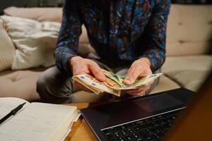 Man holding euro banknotes, sitting on sofa in front of table with laptop and notebook. Person counting remaining money after paying bills and utility expenses. Personal finance and home budget photo