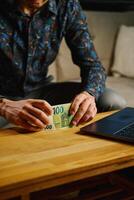 Man holding one hundred euro banknotes, sitting on sofa in front of table with open laptop. Person counting cash money at home. Personal finance and home budget concept photo