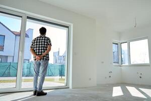 Rear view of man standing in an new empty apartment, looking out the window at modern residential neighborhood. Concept of real estate investment, property purchase and home renovation photo