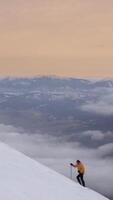 A climber in a yellow jacket makes their way up a snow-covered incline, surrounded by mist and a mountain range under a soft orange sky during twilight. Vertical footage video