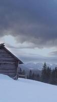 A rustic cabin sits in a snowy landscape with towering mountains in the background. Dark clouds loom overhead, hinting at a change in weather during winter. Vertical footage video