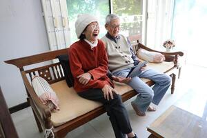 Senior asian couple are laughing and sitting on the sofa while watching funny stand up comedy show from streaming digital tablet during winter time for mental health and happiness in marriage photo