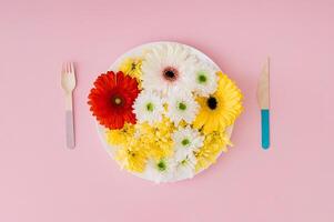 Creative trendy layout made with white plate full of colorful natural spring and summer flowers, fork and knife on pastel pink background. Flat lay style. Top view. photo