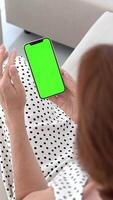 mulher sentado em sofá usando Smartphone, com vertical verde croma chave tela para brincar ou substituição, rolagem mão movimento em mostrar, casual casa contexto. video