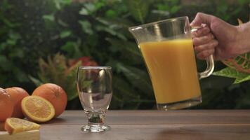 Close-up of a man's hand lifting a jug of orange juice and filling it into a glass on a wooden table against a background of green plants. video