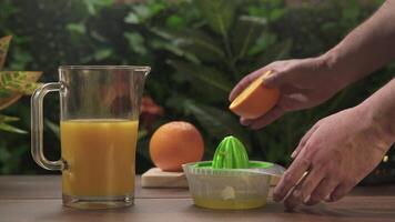 Close-up of a man's hands squeezing oranges next to a jug of orange juice on a wooden table against a background of green plants. video
