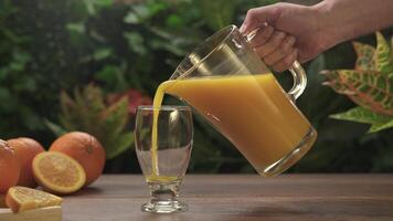 Close-up of a man's hand holding a jug of orange juice and filling it into a glass cup on a wooden table against a background of green plants. video
