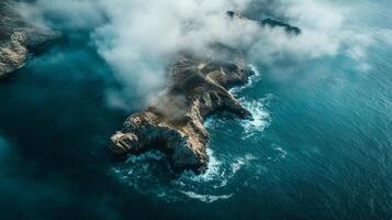 Dramatic aerial view of rocky island partially obscured by dense, low-lying fog, surrounded by deep blue ocean waters with breaking waves. photo
