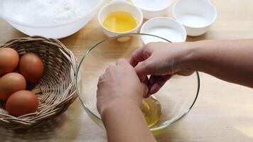 Hands Cracking Egg into Glass Bowl on Wooden Kitchen Counter with Basket of Eggs and Baking Ingredients in Background video
