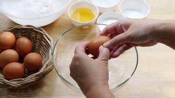 Hands Separating Egg Yolk from Egg White into Glass Bowl on Wooden Kitchen Counter with Basket of Eggs and Baking Ingredients in Background video