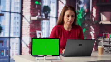 Mature woman reading forms and papers next to isolated screen and studying handwritten notes, surfing the web and staying connected for her freelance work tasks. Multitasking online. Camera A. video