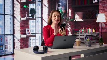 Mature adult smiling and waving during a call at home desk, showcasing modern technology and online communication in a digital and wireless lifestyle. Web conference on phone. Camera B. video
