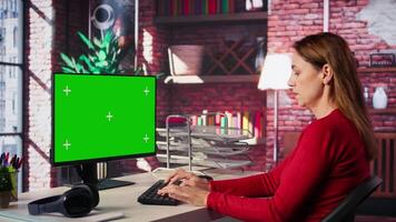 Adult woman typing on pc with chroma key screen at her loft desk, engaging in online work, multitasking efficiently and embracing the flexibility of remote tasks in a home setting. Camera B. video