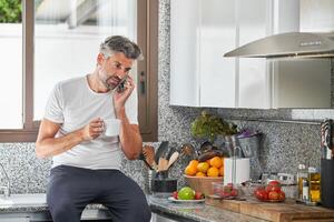 Middle aged man talking on cellphone and drinking coffee in kitc photo