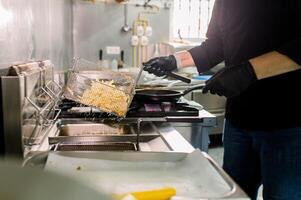 Crop anonymous male chef in black gloves with deep fryer pan preparing French fries in kitchen of restaurant with utensils photo