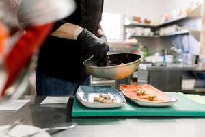 Side view of crop anonymous male cook in gloves standing at table and serving delicious broiled prawn with vegetables on plates in kitchen photo