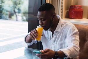 Black man browsing on phone in cafe while sipping an orange juice photo