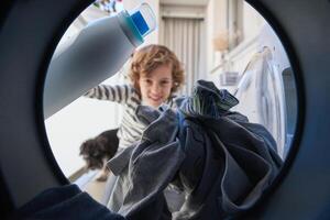 Smiling child with detergent loading clothes in washing machine photo