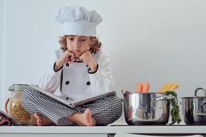 Pensive preteen boy in chef uniform sitting with opened cookbook photo