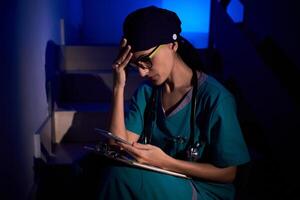 Worried woman in scrubs sitting on stairs and browsing smartphon photo