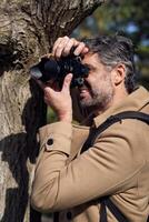 Smiling man leaning on tree and taking photo in woods