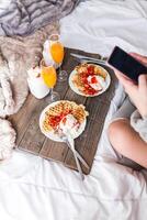 Woman with smartphone near appetizing waffles and tasty drinks on tray on bed photo
