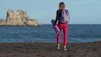 Unrecognizable female walking on black sandy beach with ocean waves, island in background during physical activity. Athlete with exercise mat and backpack. Rear view, handheld shot, real time, zoom in video