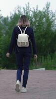 Rear view of an unrecognizable woman walking along pathway in park in summer. Model is wearing blue jacket, jeans and sneakers, is carrying small backpack on her back. Handheld, zoom in video