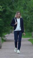 Full length view of woman walking on footpath in public park in summertime. Beautiful model dressed in blue jacket and jeans and is carrying small backpack on back. Handheld, zoom out. Vertical video